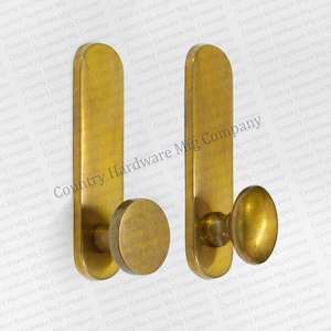 May include: Two antique brass coat hooks. Each hook features a rectangular backplate with rounded top and a circular or oval knob. The hooks are made of brass and have a vintage finish. The hooks are suitable for hanging coats, hats, or other items.
