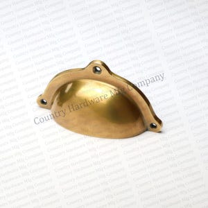 May include: A brass-colored cup pull with a curved design and three screw holes. The hardware has a smooth, reflective surface and is likely for a cabinet or drawer. The text "Country Hardware Mfg Company" is visible.