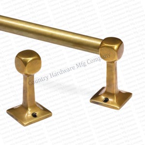 May include: A brass-toned towel bar with two matching square-based supports. The bar and supports have a vintage aesthetic, with a slightly aged finish. The supports feature a cuboid design at the top, adding a touch of classic style.