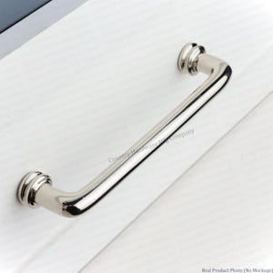 May include: A polished silver-toned metal cabinet pull handle with a smooth, rounded bar design and two circular mounting bases featuring tiered detailing, attached to a white surface with the text Country Hardware Mfg Company visible.