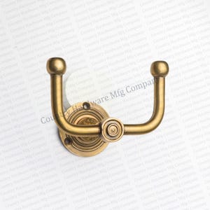 May include: A brass-colored, double hook with a circular base and decorative detailing. The hook has a curved design with rounded ends, suitable for hanging items. The hardware is likely for a coat rack or similar storage solution.