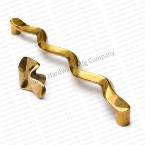 May include: A set of two brass-toned cabinet hardware pieces. One is a wavy, elongated handle with rounded ends, and the other is a matching corner piece. The hardware has a smooth, polished finish.