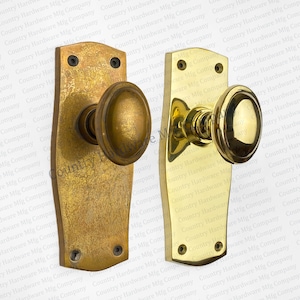 May include: Two antique brass door knobs with a rounded design. The knobs are mounted on rectangular plates with rounded edges. The plates have two screw holes each.