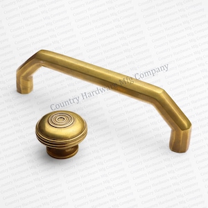 May include: A set of brass cabinet hardware, including a bar pull and a round knob. The bar pull has a geometric design, and the knob features concentric circles. Both pieces have a warm, antique finish.