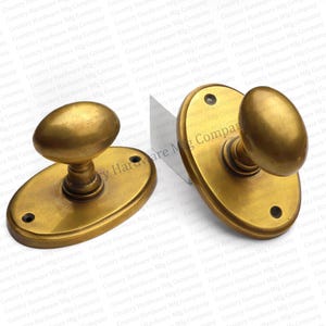 May include: Two antique brass oval door knobs. Each knob has an oval handle and a matching oval backplate with screw holes. The knobs have a warm, aged finish, suggesting vintage hardware.