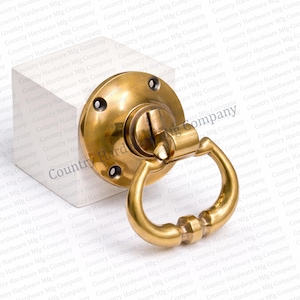 May include: A brass door pull with a circular backplate and a curved handle. The handle has a decorative design in the center. The hardware is mounted on a white surface. The text "Country Hardware Mfg Company" is visible.