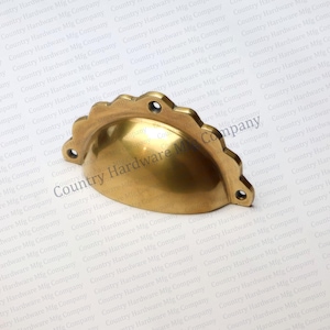 May include: A brass-colored, scalloped-edge cabinet pull with a curved, cup-like design. The hardware features three pre-drilled holes for mounting. The product is from Country Hardware Mfg Company.