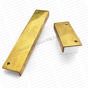 May include: Two brass-toned rectangular cabinet pulls. One is long and narrow, the other is shorter with an L-shaped design. Both have pre-drilled holes for mounting. The text "Country Hardware Mfg Company" is visible.
