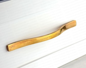 Könnte beinhalten: Ein goldfarbener Schrankgriff mit geschwungenem, gedrehtem Design. Der Griff ist rechteckig mit abgerundeten Kanten und trägt den Text "Country Hardware Mfg Company". Der Griff ist an einer weißen Schranktür montiert.