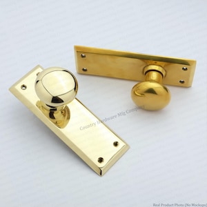 May include: Two polished brass door knobs mounted on rectangular backplates. Each plate features four small screw holes at the corners. The knobs are spherical and reflective, showcasing a smooth, metallic finish throughout the hardware set.