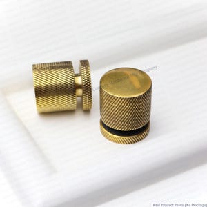 May include: Two cylindrical brass knobs featuring a textured diamond knurled pattern on the sides and a smooth, flat top surface. Each knob has a distinct circular base plate and a central stem, showcasing a metallic gold finish throughout.