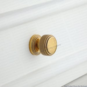 May include: A brass cabinet knob with a textured, knurled grip and a smooth, round face. The knob is mounted on a white surface, showcasing its antique finish. The knob is from Country Hardware Mfg Company.