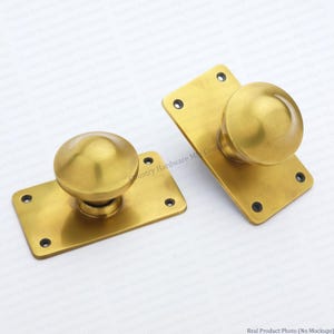May include: Two brass door knobs with matching rectangular backplates. Each knob features a smooth, spherical design attached to a flat, polished brass plate with four pre-drilled screw holes at the corners for mounting to a door surface.