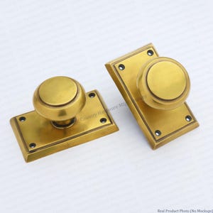 May include: Two brass door knobs with matching rectangular backplates. Each knob is spherical with a central ridge, mounted on a beveled rectangular plate featuring four small screw holes at the corners for secure installation on a door surface.