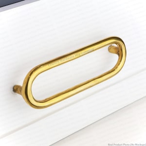 May include: A polished gold-tone metal drawer pull with an elongated oval shape. The handle features a smooth, rounded profile and is mounted onto a white surface. The text Country Hardware Mfg Company is engraved on the top of the metal handle.