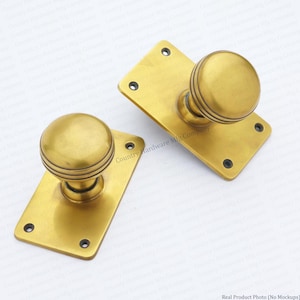 May include: Two brass door knobs with matching rectangular backplates. Each knob features a spherical shape with three horizontal grooves around the centre. The rectangular plates have four small screw holes at the corners for mounting purposes.