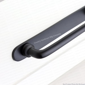 K&ouml;nnte beinhalten: Ein T&uuml;rgriff aus mattschwarzem Metall mit einer langen, flachen, rechteckigen R&uuml;ckplatte mit abgerundeten Enden und einem zylindrischen Griff. Der Text Country Hardware Mfg Company ist in Wei&szlig; auf der R&uuml;ckplatte aufgedruckt.