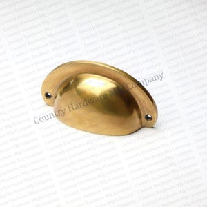 May include: A solid brass cup pull with a curved design. The hardware has two mounting holes and a polished finish. The product is from Country Hardware Mfg Company.