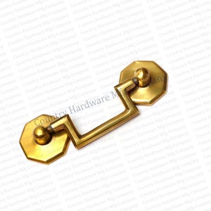 May include: A brass-toned cabinet pull with an octagonal backplate and handle. The handle has a geometric design and a rounded knob at each end. The hardware is likely for furniture or cabinetry.