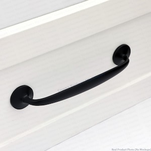 May include: A matte black cabinet pull with a curved, arching handle design and two circular mounting bases. The hardware is attached to a white, paneled surface. The text Country Hardware Mfg Company is visible on the white surface.
