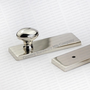 May include: A set of polished nickel hardware components, including a knob and rectangular plates. The knob has an oval top and a decorative base. The text "Country Hardware Mfg Company" is visible on the plates.