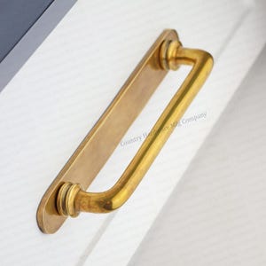 May include: A brass-toned cabinet pull with a rectangular backplate and a curved handle. The handle is a rounded rectangle shape. The text "Country Hardware Mfg Company" is printed on the backplate.