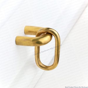 May include: A gold-toned metal cabinet pull featuring a U-shaped base attached to a vertical oval ring. The hardware has a smooth, polished finish with a reflective surface. The design consists of two distinct connected components in a metallic gold color.