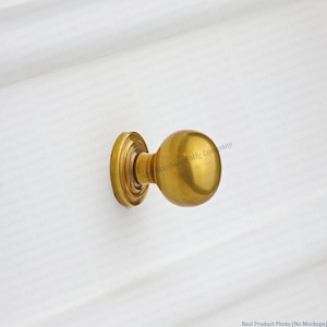 May include: A brass door knob with a round, ball-shaped handle and a circular backplate. The knob is mounted on a white surface. The hardware has a vintage or antique aesthetic.
