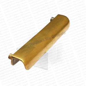May include: A brass-colored cabinet pull with a rectangular body and curved edges. The hardware features a small, cylindrical post on one end and a curved, open design on the other. The text "Country Hardware Mfg Company" is visible.