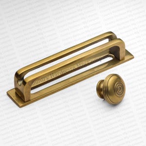May include: A set of antique brass hardware, including a rectangular pull with a curved handle and a round knob with concentric circle detailing. The hardware is likely for cabinets or drawers.