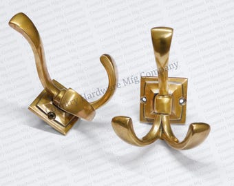 May include: Two brass-colored coat hooks. One hook has a single curved arm, and the other has three curved arms. Both hooks are mounted on square bases with screw holes. The hooks are likely for hanging coats or other items.