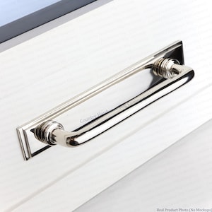 May include: A polished nickel cabinet pull featuring a rectangular backplate with beveled edges and a curved handle. The handle is attached to the backplate with two circular, ribbed mounts. The hardware has a reflective, metallic silver finish.