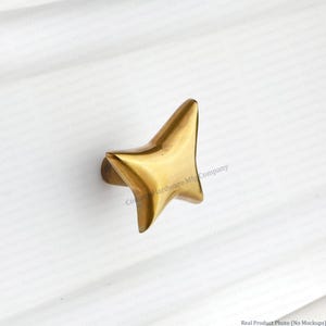 May include: A gold-toned metal cabinet knob shaped like a four-pointed star with smooth, curved edges. The star-shaped hardware features a polished, reflective surface and is mounted on a white, horizontally paneled surface.