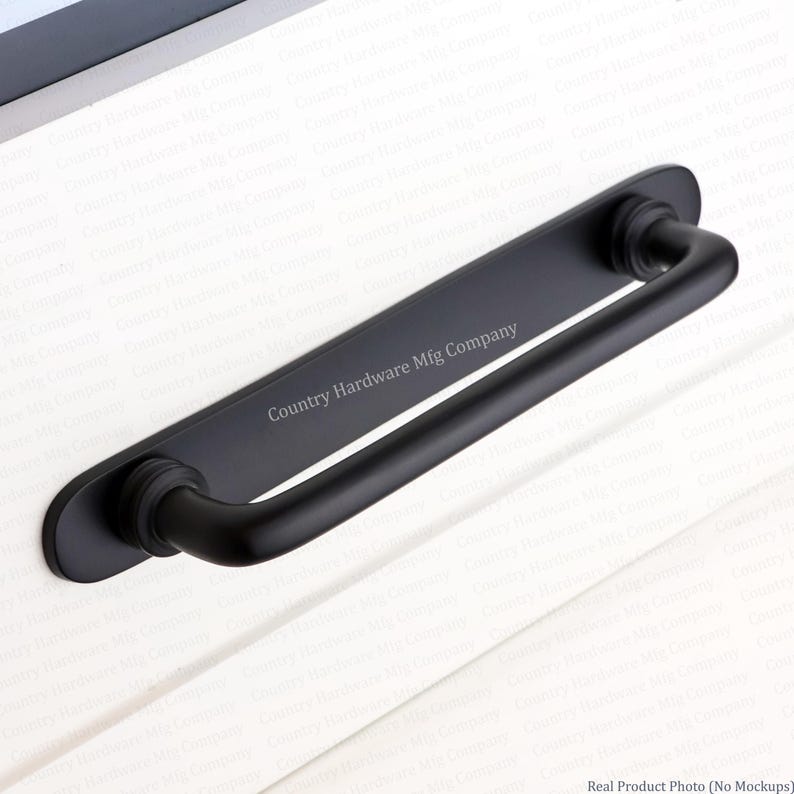 K&ouml;nnte beinhalten: Ein mattschwarzer Schrankgriff mit einer langen, rechteckigen R&uuml;ckplatte mit abgerundeten Enden und einem stabilen, zylindrischen Griff. Der Text Country Hardware Mfg Company ist in die Mitte der R&uuml;ckplatte eingraviert, die auf einer wei&szlig;en Fl&auml;che montiert ist.