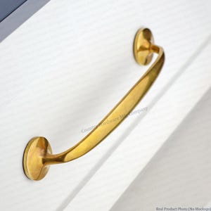 May include: A curved brass cabinet pull with a polished finish. The handle has a rounded shape and is attached to the cabinet with two circular mounting plates. The hardware is designed for easy installation and adds a touch of elegance to any furniture piece.