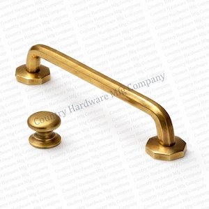 May include: A set of brass-toned cabinet hardware, including a pull handle and a knob. The pull handle is a curved bar with octagonal mounting plates. The knob has a rounded top and a stepped base.