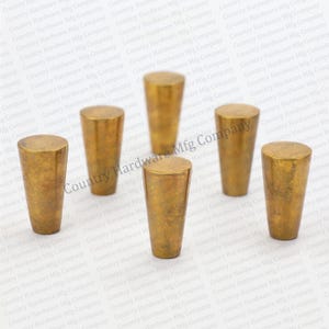 May include: Six brass-colored cabinet knobs with a cylindrical shape and a flat top. The knobs have a textured, aged appearance, suggesting a vintage or antique style. They are arranged on a white background.