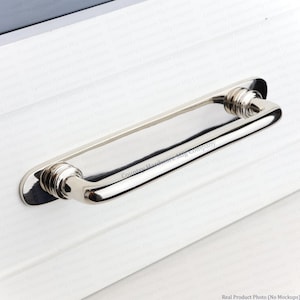 May include: A polished nickel cabinet pull with a sleek, elongated handle and two circular mounting plates. The hardware features a smooth, reflective metallic finish and a curved design that connects to the base with ribbed detailing.