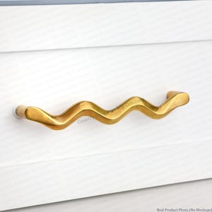 May include: A brass-colored cabinet pull with a wavy, organic design. The handle is mounted on a white cabinet door. The pull has a smooth, polished finish and a modern aesthetic, suitable for drawers or cabinet doors.