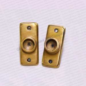 May include: Two brass curtain rod finials with a round, slightly domed top and a small hole in the center. The finials are rectangular and have a smooth, polished finish.