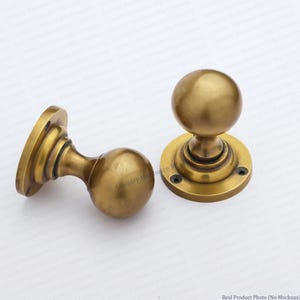 May include: Two brass door knobs with a spherical handle and a circular mounting plate. Each knob features a smooth, polished metallic finish with a warm golden tone. The mounting plates have visible screw holes for installation on doors.