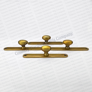 May include: Three gold-toned metal cabinet door pulls with a rounded knob design. Each pull is mounted on a rectangular metal plate. The plates are attached to a wooden door.