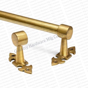 May include: A brass-colored curtain rod set, featuring a long rod and two decorative brackets. The brackets have ornate, stylized designs and are mounted on a white background. The set is designed for window treatments.