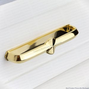 May include: A polished gold-tone metal drawer pull with a recessed center grip and a central decorative protrusion. The surface of the hardware features the text Country Hardware Mfg Company printed in a small, repeating pattern across the metal.