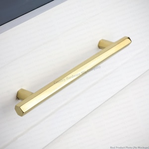May include: A gold-toned metal cabinet pull with a hexagonal profile and a smooth, polished finish. The handle is mounted horizontally on a white surface, featuring a clean, geometric design with faceted edges and two mounting points.