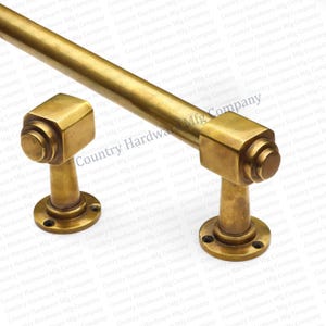 May include: A brass-toned towel bar with two matching wall mounts. The bar is cylindrical, and the mounts have a square design with a circular accent. The hardware is designed for bathroom or kitchen use.