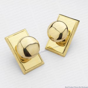 May include: Two polished brass door knobs, each featuring a spherical handle mounted on a rectangular backplate with beveled edges. The metal surface has a reflective, smooth finish, showcasing a classic design for interior or exterior doors.