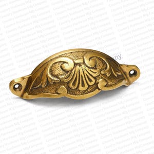 May include: Ornate brass-toned cabinet pull with a shell and scroll design. The hardware features two mounting holes and a curved handle, perfect for adding a vintage touch to furniture. The pull measures approximately 5 inches in length.