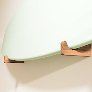 May include: A light green surfboard mounted on a wall with two wooden brackets. The brackets are a light brown color and are designed to hold the surfboard securely. The surfboard is curved and has a smooth surface.
