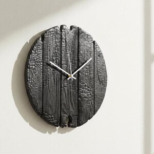 May include: A round, black wooden wall clock with white hands. The clock face is made of vertical wooden planks, giving it a rustic appearance. The clock is mounted on a white wall.